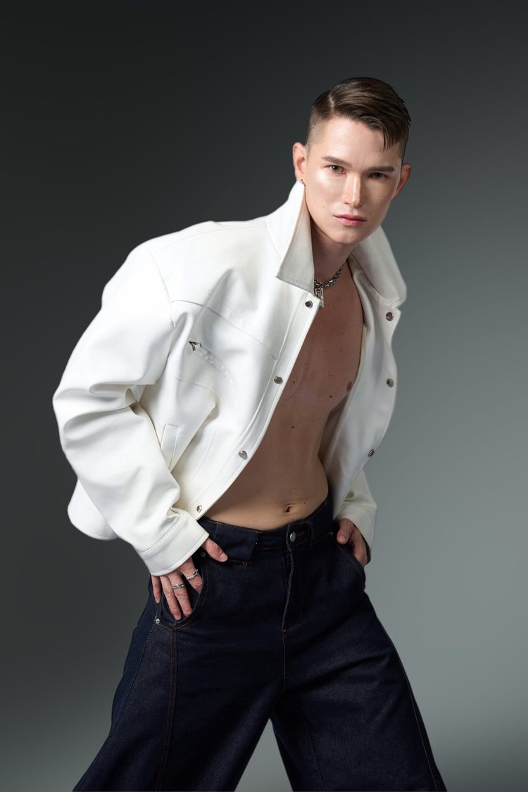 Dee Mak in an open white leather jacket and dark wide-leg denim, chain necklace, turned toward camera against a dark studio backdrop