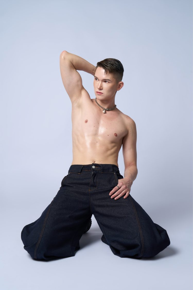 Dee Mak shirtless kneeling with one arm raised behind his head, silver chain necklace, dark wide-leg denim