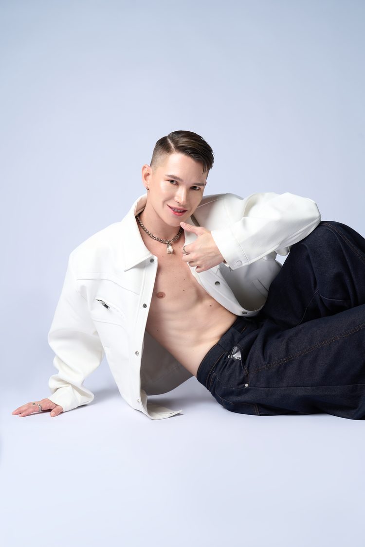 Dee Mak seated on the floor, shirtless in an open white leather jacket, smiling with hand near his chin